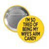 Yellow pinback button with text 'I'm so tired of being my wife's arm candy' printed in black.