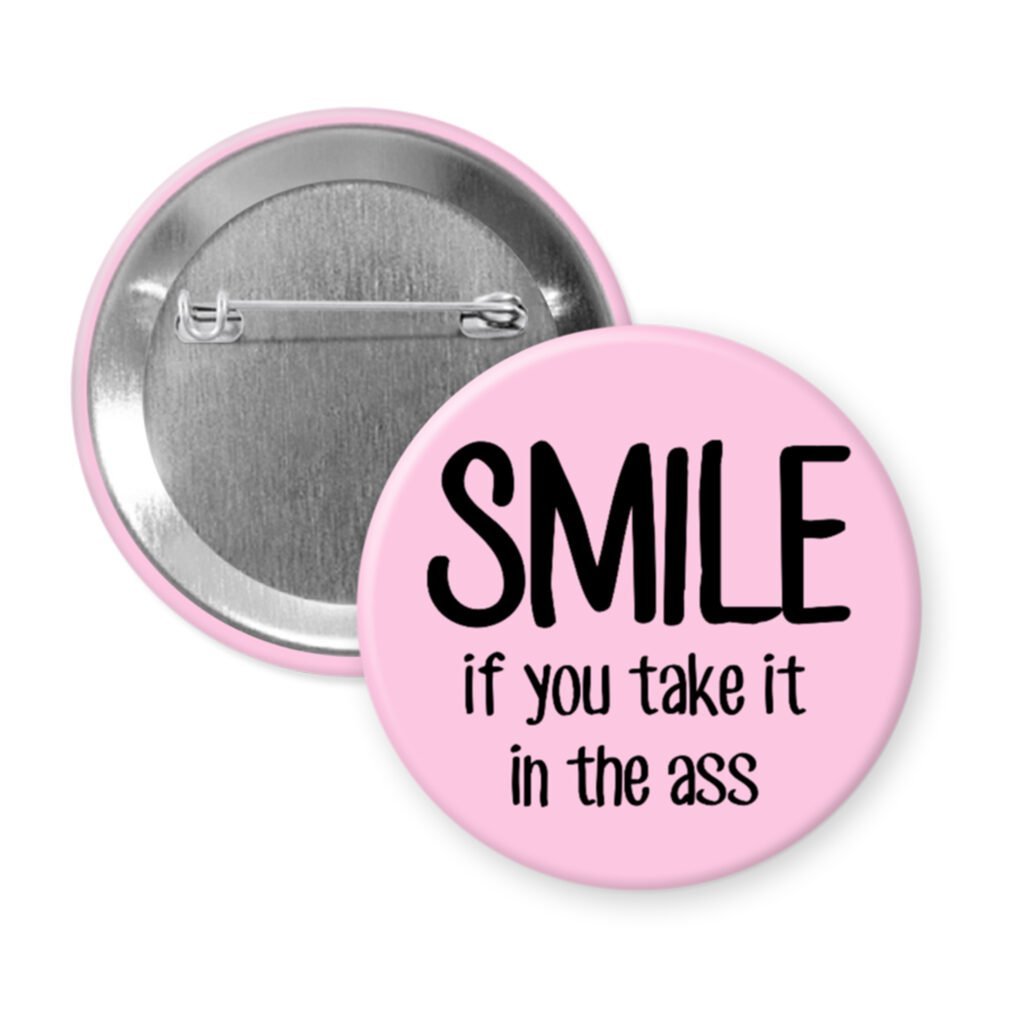 Pink pinback button with the text 'SMILE if you take it in the ass' printed in black.