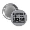 Grey pinback button with bold black text reading ‘Adulting is a Scam’ in mixed block and distressed fonts. The design includes a smiley face, star, and flower icons, with a checkerboard stripe along the bottom.
