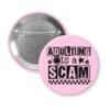Pink pinback button with bold black text reading ‘Adulting is a Scam’ in mixed block and distressed fonts. The design includes a smiley face, star, and flower icons, with a checkerboard stripe along the bottom.