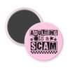 Pink magnet with bold black text reading ‘Adulting is a Scam’ in mixed block and distressed fonts. The design includes a smiley face, star, and flower icons, with a checkerboard stripe along the bottom.