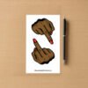 Sticker sheet with two brown hand designs showing middle‑finger gesture and red nails, placed beside a pen on wood surface.