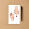 Sticker sheet with two light‑skin hand designs showing middle‑finger gesture and red nails, placed beside a pen on wood surface.