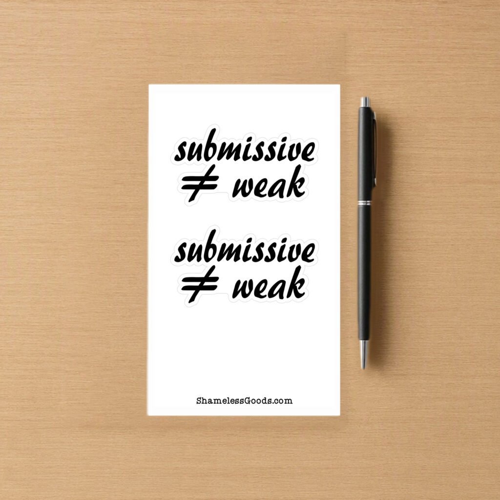 Sticker sheet on a wooden surface beside a black pen, featuring two identical black script phrases that read submissive ≠ weak, each outlined in white. Branding at bottom reads ShamelessGoods.com.