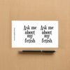 Sticker sheet on a wooden surface beside a black pen, featuring two identical black text designs that read Ask me about my fetish in bold serif font. Vertical branding text on the left reads ShamelessGoods.com.