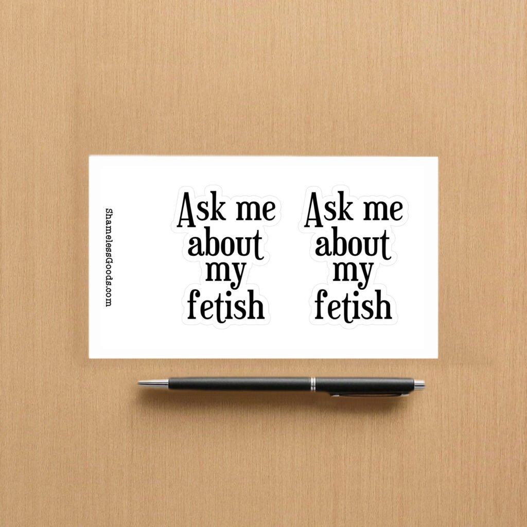 Sticker sheet on a wooden surface beside a black pen, featuring two identical black text designs that read Ask me about my fetish in bold serif font. Vertical branding text on the left reads ShamelessGoods.com.
