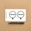 Sticker sheet on a wooden surface beside a black pen, featuring two identical circular chain designs with padlocks at the bottom and the word OWNED in bold black letters inside each circle. Vertical branding text on the left reads ShamelessGoods.com.