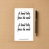 Sticker sheet on a wooden surface beside a black pen, featuring two identical black script phrases that read A bound body frees the mind, each outlined in white. Branding at bottom reads ShamelessGoods.com.