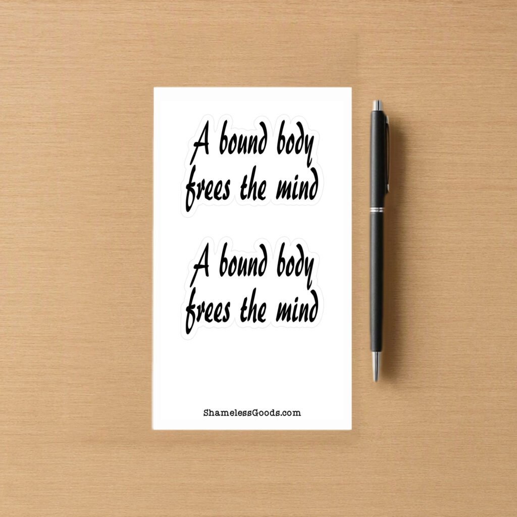 Sticker sheet on a wooden surface beside a black pen, featuring two identical black script phrases that read A bound body frees the mind, each outlined in white. Branding at bottom reads ShamelessGoods.com.